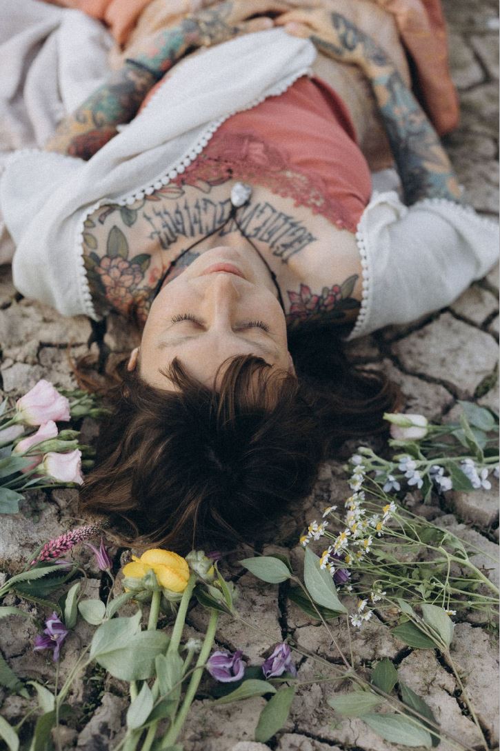 Angelina Katharina Petersen in Leipzig, liegend auf rissiger Erde umgeben von Blumen, mit geschlossenen Augen und Tattoos, friedliche Ausstrahlung