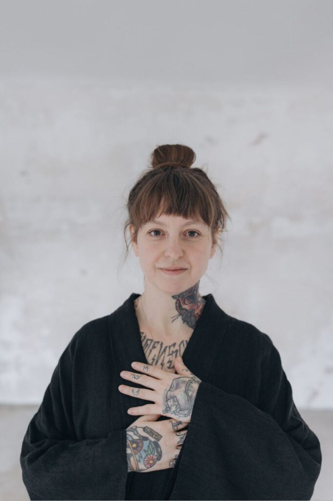 Angelina Katharina Petersen, schaut in die Kamera, trägt einen schwarzen Kimono, hat die Hände auf ihrem Herzen liegen. Sie ist Meditations und Yoga Lehrerin in Leipzig
